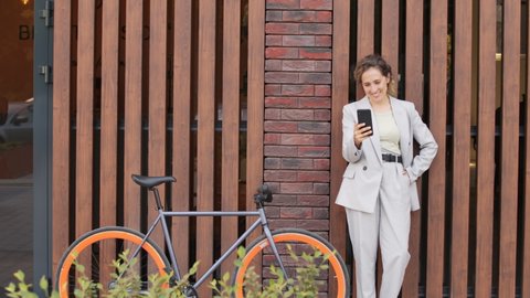 Front-view stab slowmo shot of smiling young elegant businesswoman in pant suit having video chat on smartphone standing against wall outdoors next to her bike