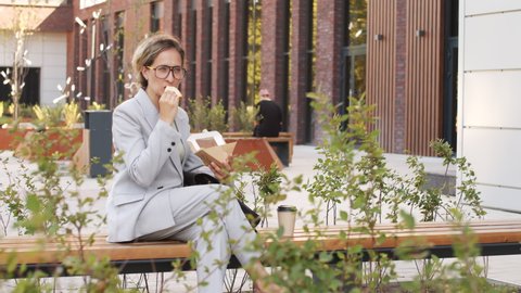 Medium slowmo of young Caucasian businesswoman in elegant pant suit and eyeglasses eating sandwich outdoors sitting on bench near modern office buildings
