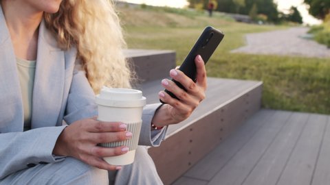 Midsection slowmo of unrecognizable businesswoman in grey pant suit holding reusable coffee cup while scrolling through smartphone sitting outdoors in park