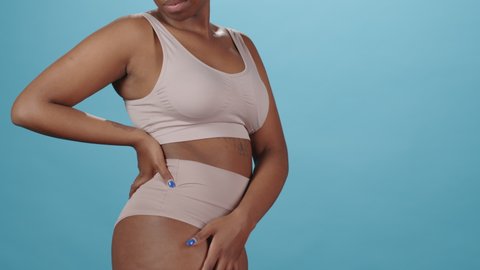 Cropped medium slowmo portrait of young plus size African-American woman in underwear showing her natural healthy body standing on blue background in studio