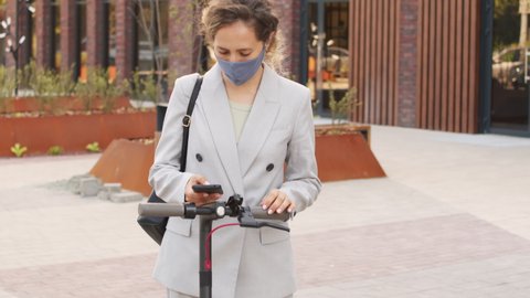 Tilt-up slowmo shot of elegant young businesswoman in face mask activating electric scooter with app on her smartphone and starting ride outdoors in city downtown