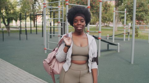 Medium slowmo portrait of smiling African-American woman with pink sports bag looking at camera standing at outdoor sports ground with exercise equipment in background