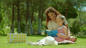 Mother and her little daughter are having a picnic outdoors on the grass. Little daughter is reading a story from a book to her mother. Mothers Day. Slow motion video.