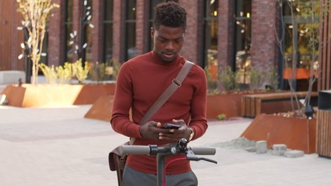 Tilt-up medium slowmo shot of young handsome African-American man in smart casualwear setting up electric kick scooter using app on his smartphone standing outdoors in business district