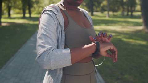 Midsection slowmo shot of unrecognizable African-American woman in sportswear setting up smartwatch on her wrist while jogging outdoors in park on summer day