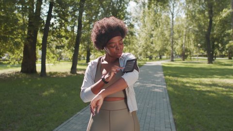 Medium slowmo shot of young African-American woman in sportswear using smartphone attached to armband while having outdoor sports training in park on summer day