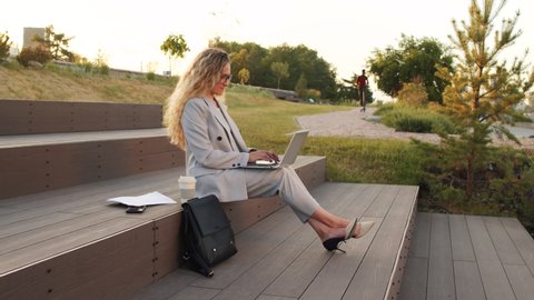 Side-view stab shot with slowmo of young confident business woman in grey pant suit and eyeglasses working on laptop and drinking coffee from plastic cup, sitting on stairs in park at sunset