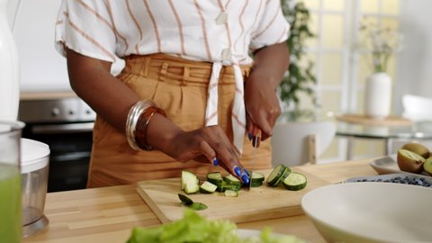 Midsection slowmo shot of unrecognizable hands of African-American woman cutting fresh cucumbers on wooden cutting board at kitchen