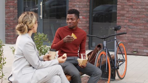 Medium slowmo shot of young interracial business couple having conversation while eating sandwiches and drinking coffee sitting on bench outside modern office building during lunch break