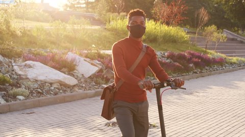 Medium slowmo portrait of young African-American businessman in red turtleneck and face mask posing with electric kick scooter standing in urban park on sunny day