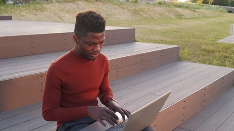 Medium slowmo shot of young African-American businessman in smart casualwear writing e-mails or working on laptop computer sitting outdoors in park on summer evening