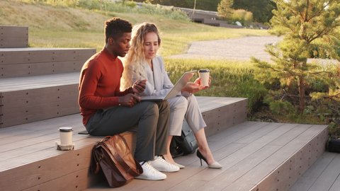 Slowmo stab shot of young interracial business couple in smart casualwear looking at laptop and having conversation sitting with take away coffee on stairs in park on sunny day