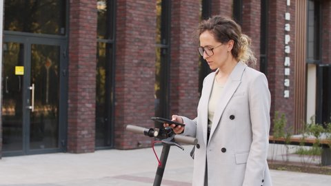 Tilt-up slowmo shot of confident Caucasian businesswoman in elegant pant suit using electric scooter for transportation in city downtown in summer