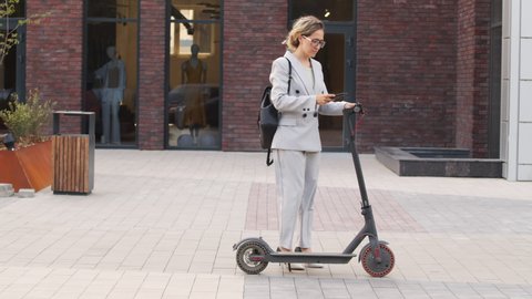 Slowmo stab shot of young Caucasian businesswoman in elegant pant suit and heels standing on electric kick scooter starting ride outdoors in city downtown on sunny day