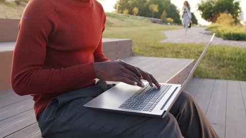 Midsection slowmo shot of hands of unrecognizable African-American businessman in smart casualwear typing on laptop keyboard, sitting on stairs in park in summer