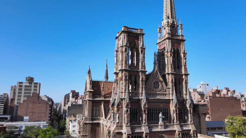 Aerial pan shot capturing details facade of gothic style church of capuchinos, Sacred Heart of Jesus with birds flying pass the building against clear blue sky in downtown Cordoba city, Argentina.