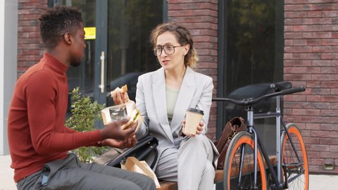 Medium slowmo shot of contemporary multi-ethnic business couple having lunch outdoors together, chatting while eating sandwiches sitting on bench in city downtown