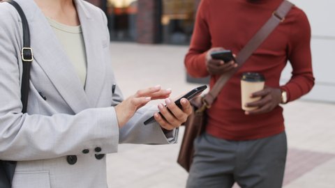 Midsection slowmo shot of unrecognizable business man and woman in smart casualwear scrolling through smartphones standing outdoors in city downtown