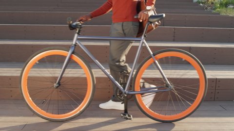 Cropped slowmo stab shot of unrecognizable African-American man in smart casualwear walking with bike outdoors on sunny day