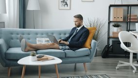 Young man wearing jacket and home shorts is talking during online presentation sitting on couch in modern apartment using modern laptop.