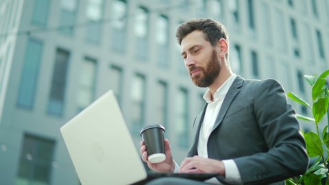 Caucasian bearded business man freelancer working on break with laptop and cup of coffee outdoors. Male office worker sitting on a bench on a city street on a modern urban background Workplace outside