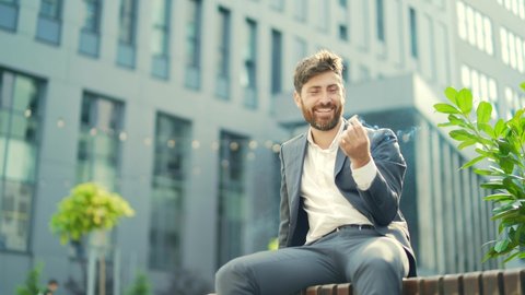 Caucasian bearded business man smoking cannabis outdoors sitting on city park bench on urban street background. Male employee Businessman. Office worker in suit Relieves stress a marijuana outside