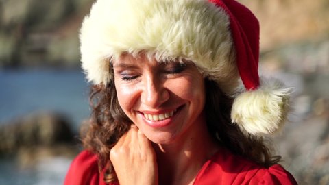 Portrait of a woman with makeup smiling at the camera and playing in a Santa hat on the beach, seashore. 4K