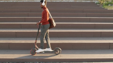 Slowmo stab shot of young African-American man in smart casualwear and headphones riding electric scooter along city on warm sunny day