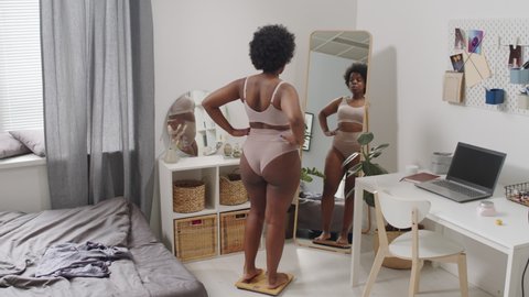 Rear-view full-length slowmo shot of young plus size African-American woman in underwear standing on scales at bedroom and examining her body in mirror