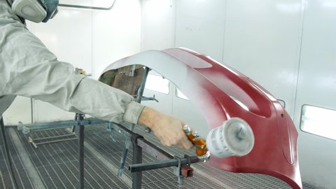Man working in car service dressed in special uniform and mask paints white auto bumper red using airless spray gun in booth closeup