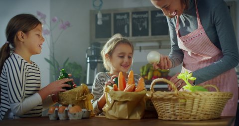 Cinematic shot of happy little daughters helping to their mother to put in basket fresh vegetables from eco-friendly paper bags delivered by courier to home after making online shopping grocery order.