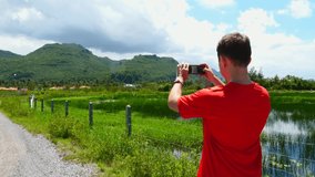 Young man shooting landscape video, photos for social media on smartphone, zooming camera gesture. Male making horizontal panorama clip, pictures of green tropical mountains on mobile phone. Back view