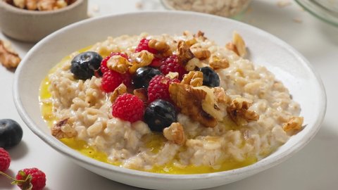 Pouring honey in oatmel porridge with fresh berries, nuts and butter in a plate. Healthy breakfast. Vegan food. 4k video