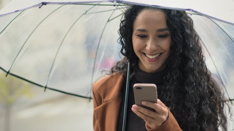 Portrait young beautiful carefree happy hispanic woman girl with umbrella stands in city rainy weather smiling typing on mobile phone chatting gets good message in smartphone waiting looking to side