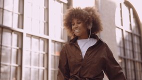 Medium Slow Motion Tracking Shot Of Young Woman With Afro Listening To Music Through Headphones, Smiling And Dancing Down Street