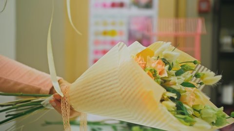 Female florist makes flower bunch, she cuts off wrapping paper working in flower shop. Cute young professional makes bouquet in floral store.
