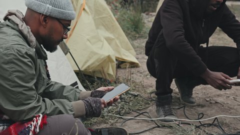 Tracking shot of unrecognizable refugees charging and using their mobile phones sitting on their laps next to extension cord outdoors at tent city
