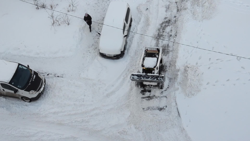 Skid steer loader removes snow from the city streets. Top view of the road with cars and snow blower. Seasonal work in winter snowy city. Equipment and city worker. 