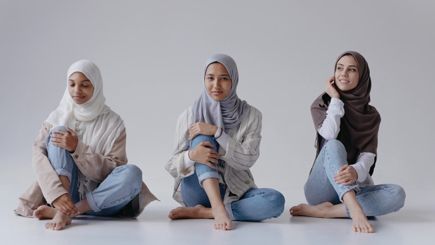 Three muslim girls sit together smiling and laughing in bright contemporary studio. Beautiful muslim women smile at the camera. Studio with white cyclorama. High quality 4k footage