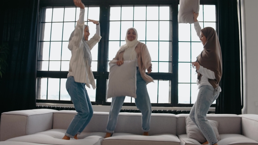 Three muslim women jump on the couch fighting with pillows, listening to music. Beautiful muslim women smile and laugh. Studio with calm and natural colors. High quality 4k footage