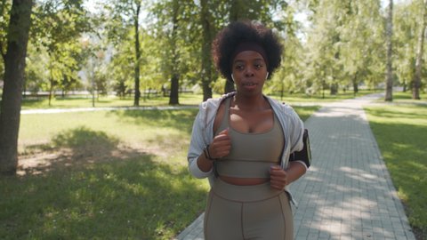 Tracking medium slowmo shot of young active African-American woman in tight sportswear, wireless earphones and smartphone in armband jogging alone in park on sunny day