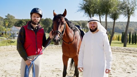 Islamic sheikh owner of the horse standing at the desert place with male jokey and smiling to the camera during the sunny day. Rich people hobbies concept