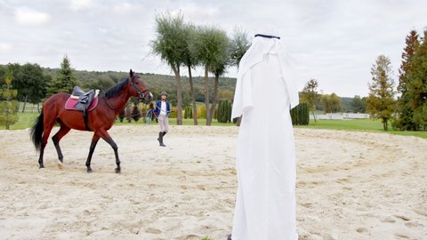 Confident arabian sheikh standing at the grass and looking at the caucasian woman training his expensive horse. Rich people concept