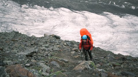 A woman in the mountains climbs up with a large backpack with equipment and trekking poles. Climber in the rocks walks up the trail during the hike