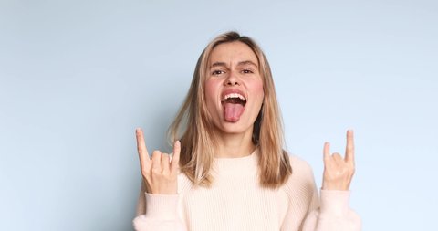 Carefree delighted beautiful girl demonstrating tongue out and rock and roll hand gesture, punk sign, crazy about success. Happy cute young girl makes rock n roll sign isolated on blue background.