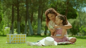Mother and her little daughter are having a picnic outdoors on the grass. Mother watching video on her smartphone with her little daughter. Mothers Day. Slow motion video.