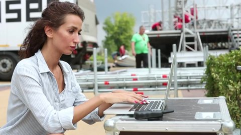 Installation of stage equipment and preparing for a live concert open air. Event manager portrait. Summer music city festival. Young serious woman work with her laptop near the stage.