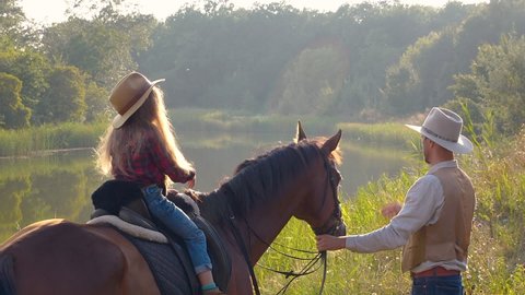 Cowboy and his daughter on horseback on the river bank. Happy cowboy family. Slow motion.