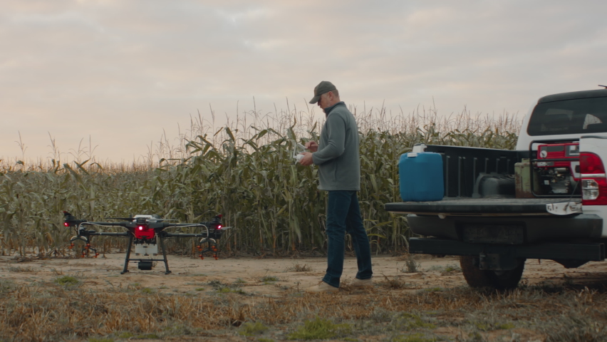 Farmer controlling a huge intelligent agriculture drone with spray nozzles near corn field early in the morning