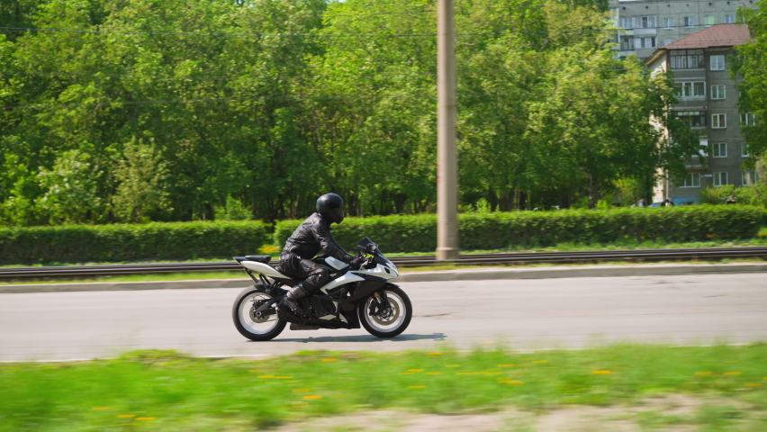 Skilled rider in black leather uniform and helmet drives powerful bike along large road past green park in city on sunny day side view slow motion